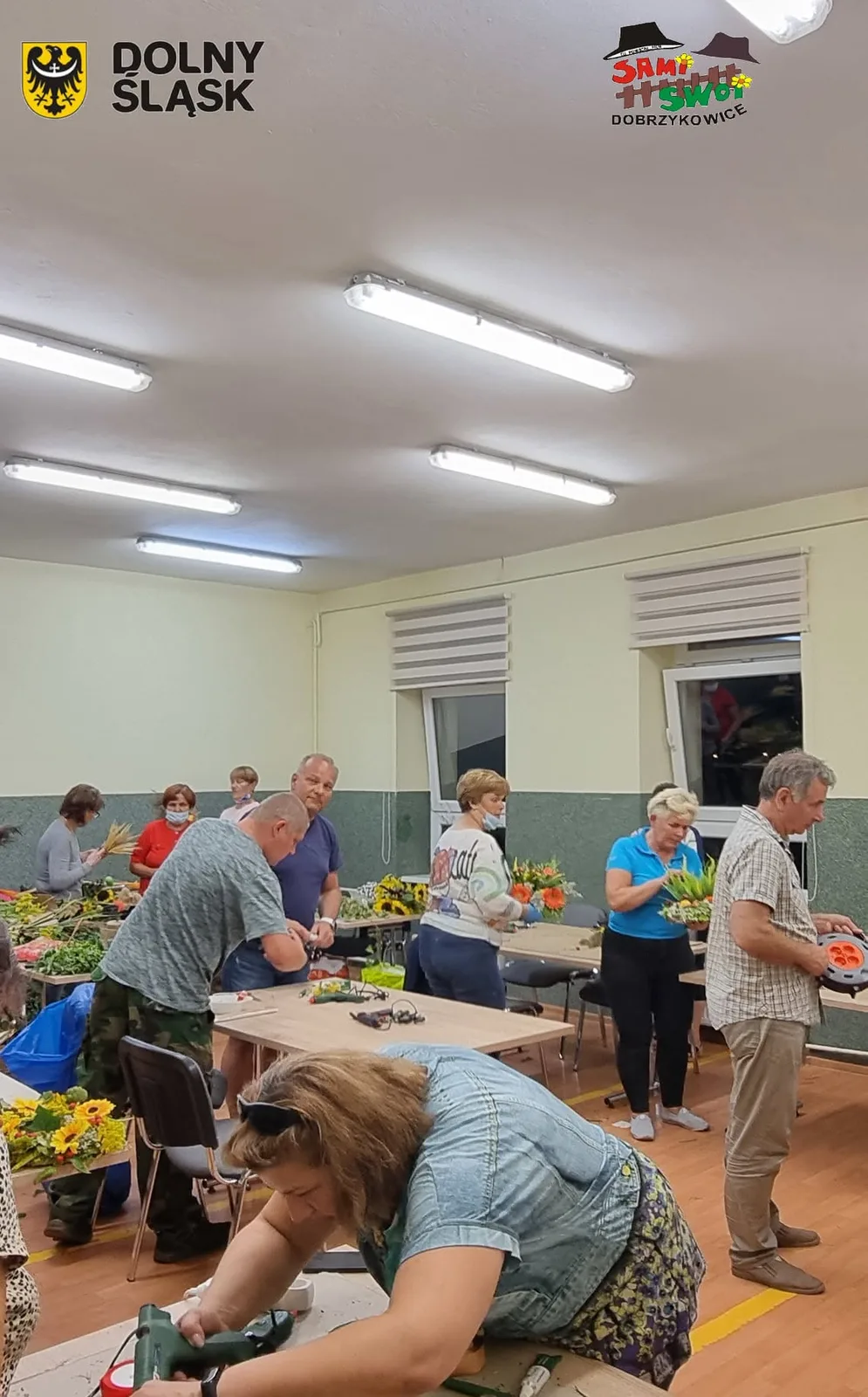 Mieszkańcy tworzą kompozycje kwiatowe przy stołach w świetlicy.
